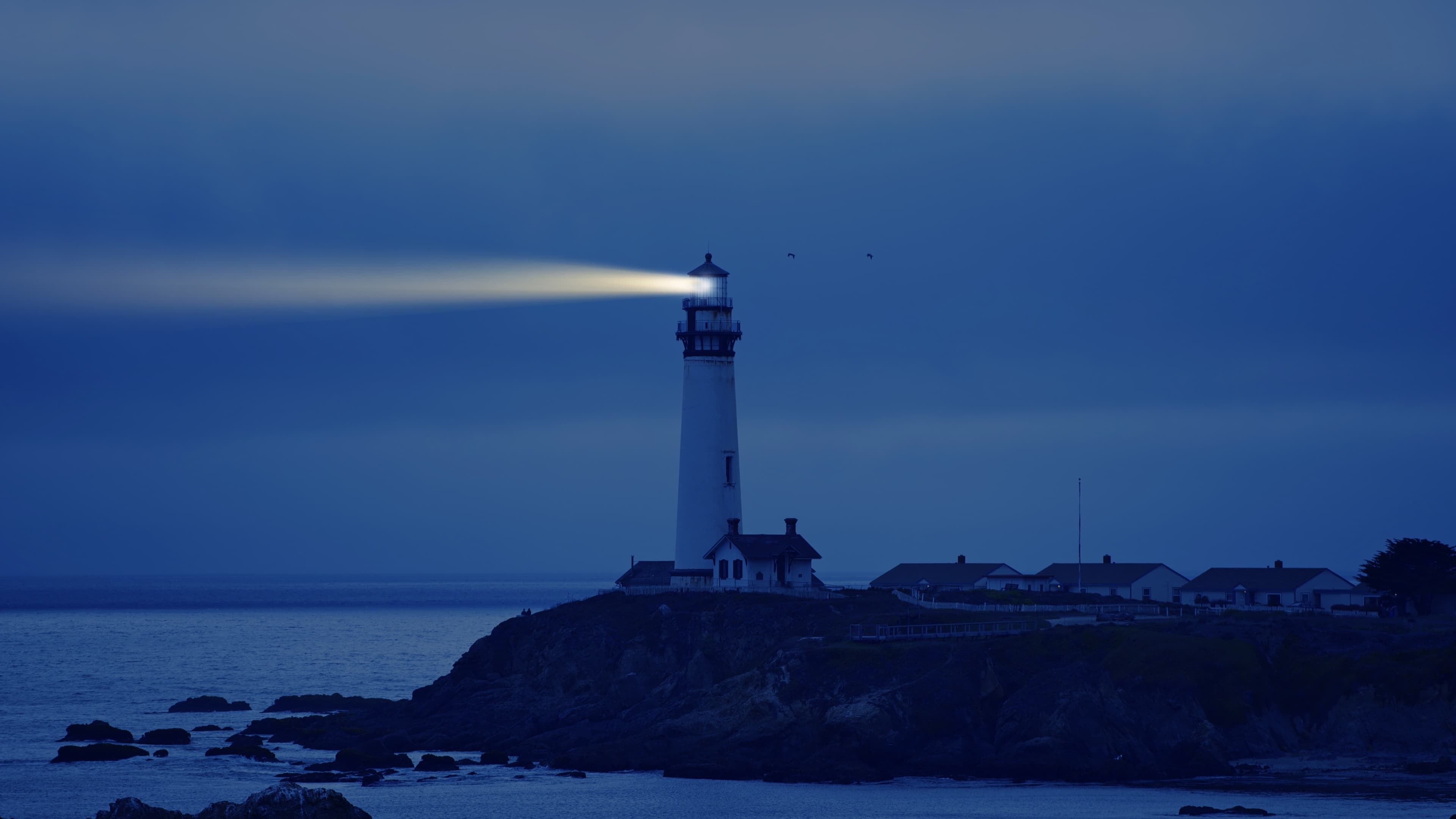 A lighthouse on a rocky shore projects its light over calm waters during an early morning darkness.